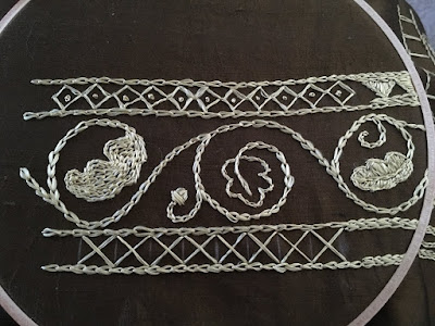 An embroidery sampler in gold thread on olive-green silk stretched in a wooden embroidery hoop, showing some narrow borders with diamond fill and large cross stitches, with a large scrolling vine down the center panel. The vine carries three oak leaves, one outlined, one filled with chain stitch, and one filled with satin stitch, and a single acorn.