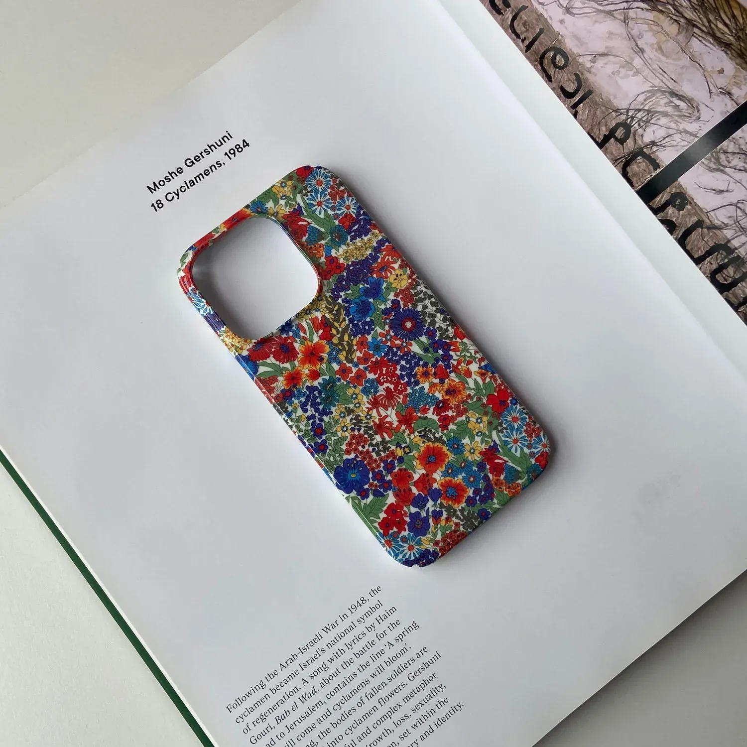 A colorful floral phone case sitting on top of a book with a painting of 18 Cyclamens by Moshe Gershuni in the background.