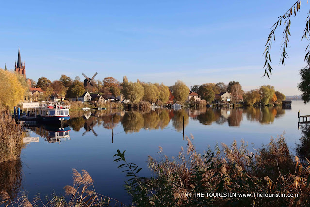 A village with a church tower a windmill and colourful houses, trees in autumn foliage, and a blue-white ferry on a pier reflected in a still lake under a soft blue sky.