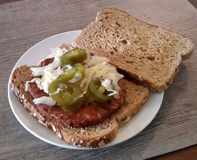 Sandwich consisting of vegetarian patty on multi-grain bread. The sandwich is displayed open, to reveal the toppings of sauerkraut and jalapenos atop the veggie patty. The bread is toasted. Sandwich is arranged on a plain white plate atop a grayish, woodgrain-patterned surface