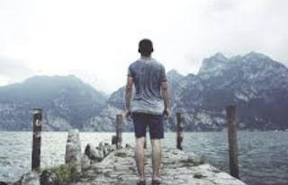A person wearing a t shirt and shorts, standing on a stone pier, looking out over a lake towards mountains