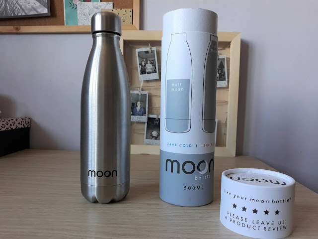 The image shows a stainless steel reusable water bottle standing next to it's white cardboard tube