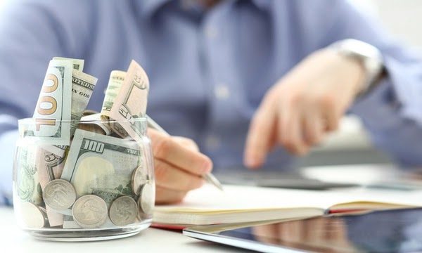 Glass jar filled with cash and coins, prominently displaying U.S. currency, with a person's hand engaged in note-taking, symbolizing financial planning for an emergency fund.