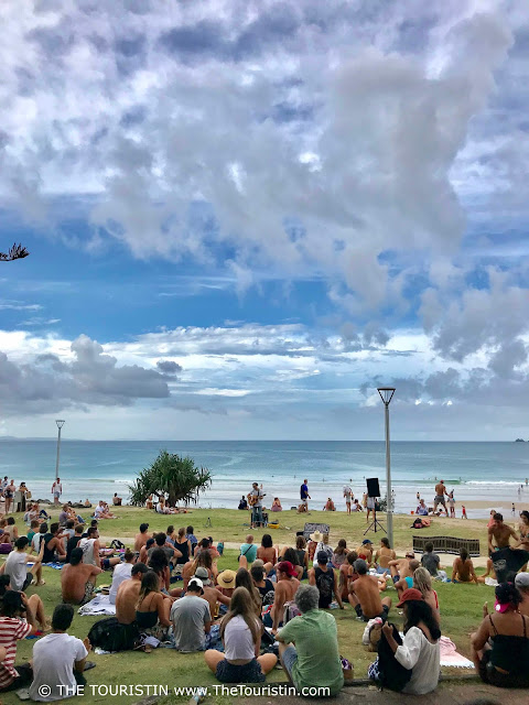 A guitar playing busker surrounded by a sitting audience und a cloudy blue sky with the green-blue coloured ocean as a background..