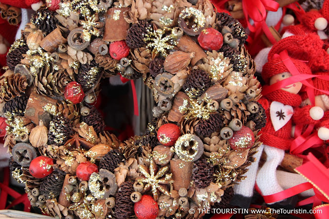 A Christmas wreath made from dried wood and pinecones sprayed partly with golden glitter stands next to a bowl with small knitted red-coloured figurines with a white felt heart on their jumpers.