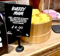 A photo of a bunch of yellow and white bee shaped soaps on a light brown shelf with a black rectangular card that says buzzy mum soap in white font on a bright background