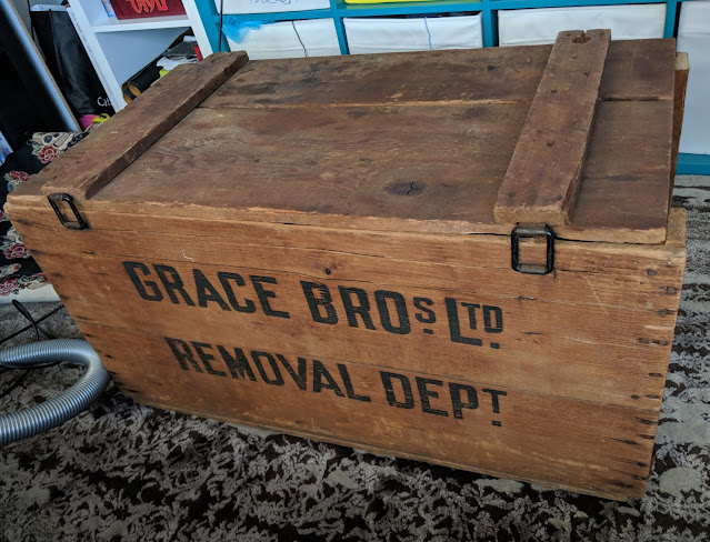 A photo of a wooden crate with a lid, with the words "Grace Bros Ltd Removal Dept" printed on the side.