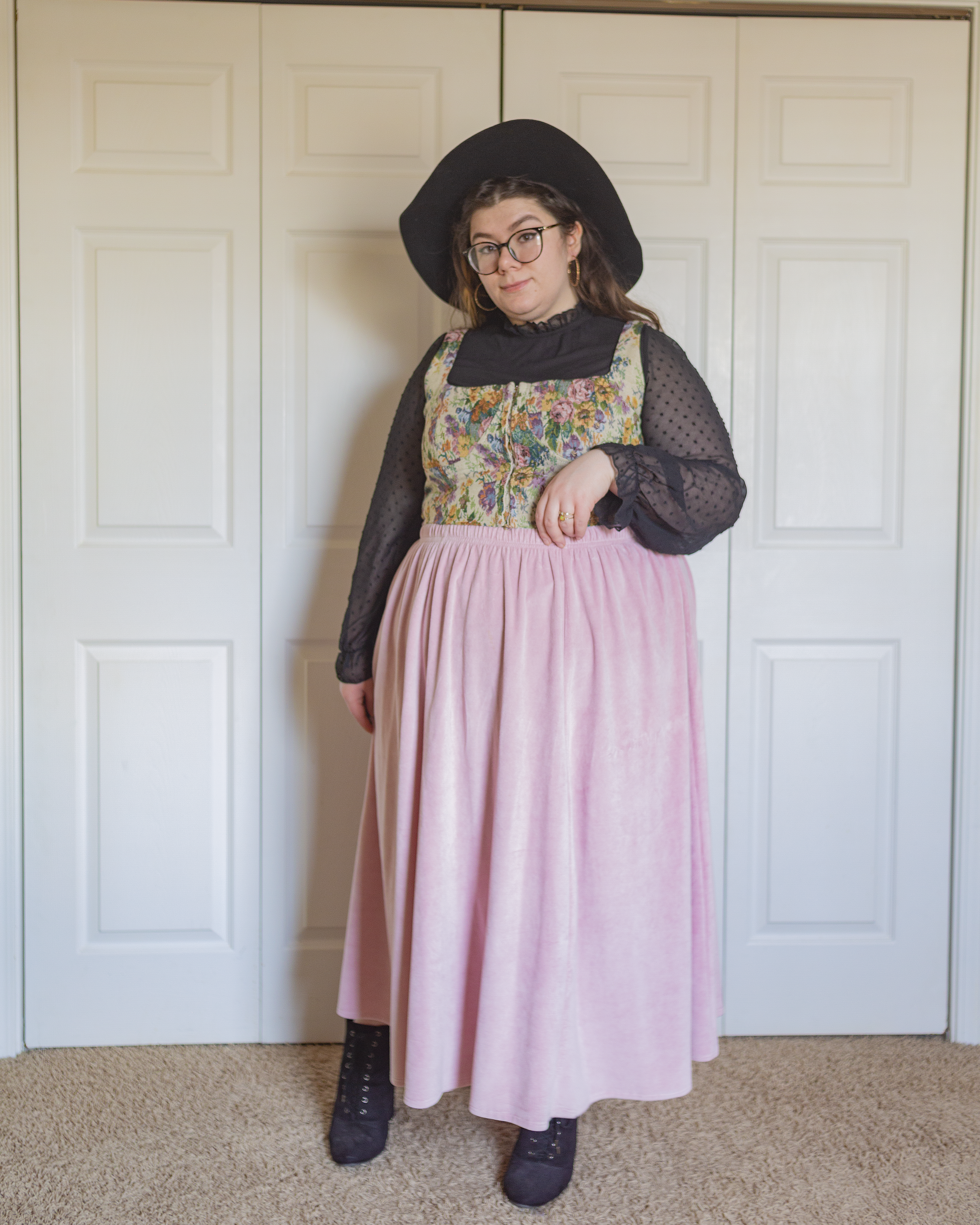 An outfit consisting of a black wide brim floppy hat, a black high neck with sheer polka dot bishop sleeves with a a cream, pastel pink, pastel blue, pastel green and lilac tapestry style corset top over and a pastel pink velour midi circle skirt and black lace up midi boots.