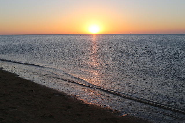 La orilla de una playa y el mar con la puesta de sol en el horizonte y su estela naranja sobre el agua.