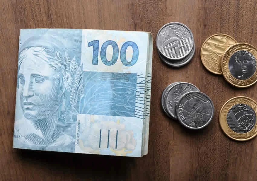 Imagem mostra uma mesa de madeira com algumas moedas e notas de 100 reais dobradas ao meio.