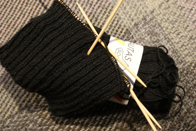 En resårstickad rudstickning i svart garn som ligger på en gråbrun vävd pläd / A circular ribbed knitting o with black yarn on a grey and brown blanket