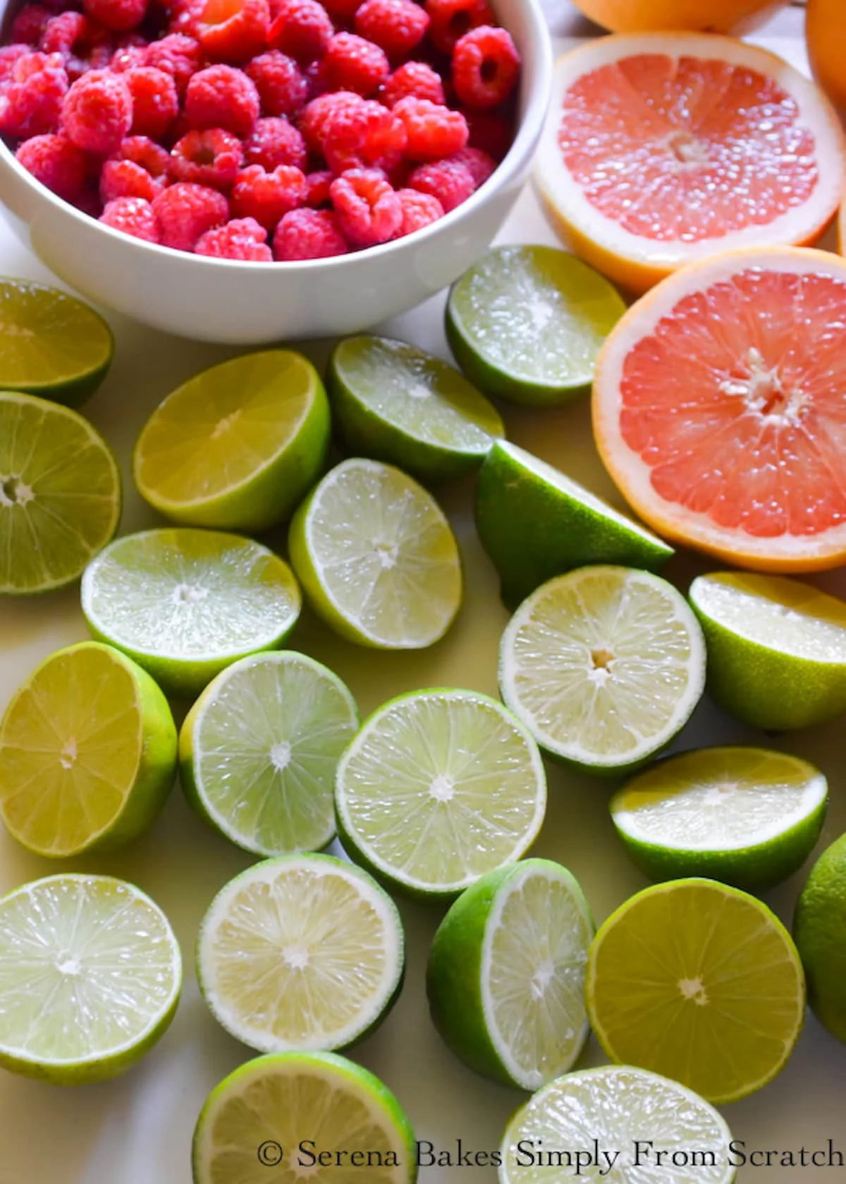 A down shot of limes sliced in half, a white bowl of fresh raspberries in the top left corner, and grapefruit sliced in half.