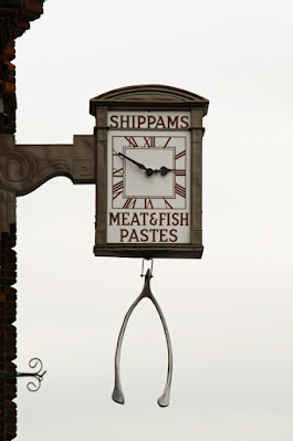 Photograph of a clock extending from a building facade on a scrolled bracket. The clock has a rectangular face with the word 'Shippams' above the dial, 'Meat & fish pastes' below. A large metal wishbone hangs beneath it.