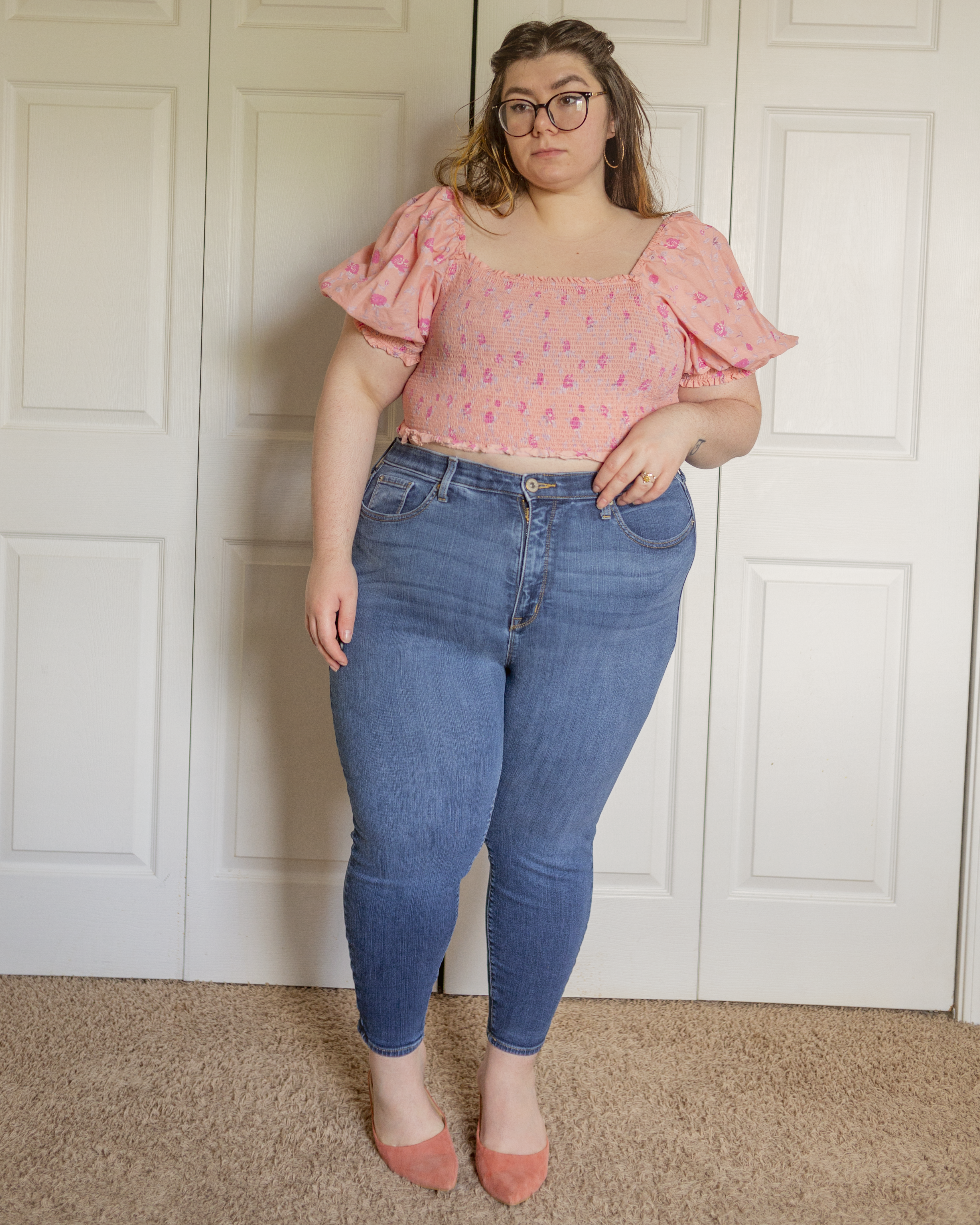 An outfit consisting of a pastel pink puffy sleeve crop top with dark pink, yellow and blue florals and medium wash high waist skinny jeans and pink suede clogs.