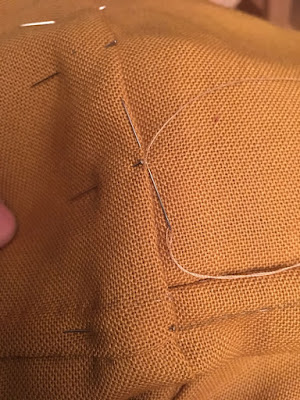 A close-up of a pinned seam in muddy-gold fabric, with a silver needle threaded with tan sewing thread inserted for the next slip stitch.