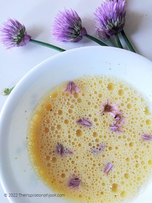 A white bowl with frothy yellow beaten eggs and purple chive blossoms and petals