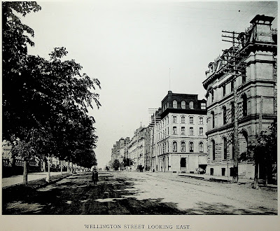 Photo taken from Wellington Street, looking east, at around O'Connor Street, with the street extending into the horizon. From left to right: The stone and wrought iron fence at the perimeter of Parliament Hill, the north sidewalk (material unclear), a boulevard planted with a continuous row of trees, the roadway which is dirt and rutted, a bicyclist in the road heading straight toward the camera, a horse drawn carriage further away on the opposite side of the street, telephone poles each with nine rows of eight insulators, the south sidewalk (concrete?) with some trees, buildings on the south side of Wellington Street, all around 4 storeys tall. The photo has an all-caps caption at the bottom (from the book from which it was scanned), Wellington Street Looking East