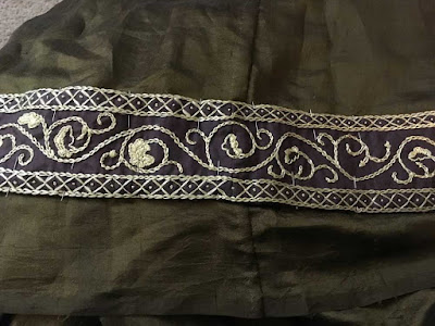 A pinned band of gold oak-leaf embroidery with narrow diamond borders on brown silk, attached to green-gold silk. The seam in the embroidered band is just visible where a diamond is distorted into a hexagon, but the seam in the green-gold base fabric is evident.