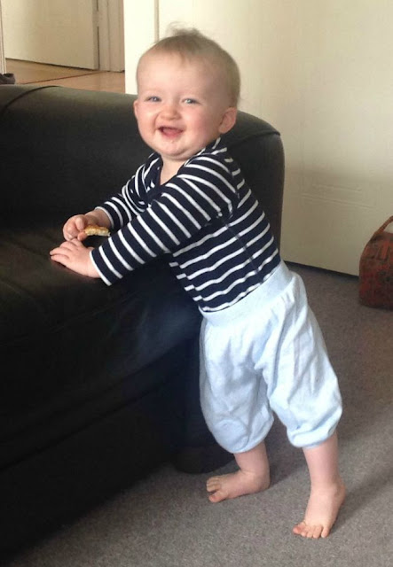 Baby standing against sofa toast in hand big smile on face