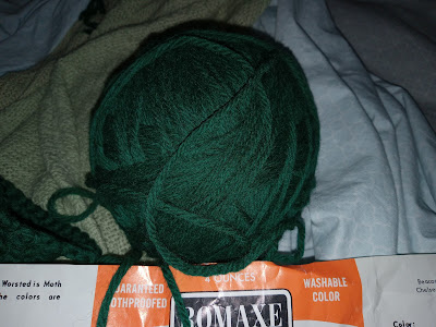 A ball of dark green yarn sits against a pale green blanket and a gray sheet with the ROMAXE label below it.