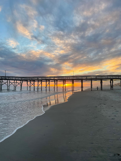 I am walking closer to the pier as I can see that the sunset is going to be visible once the sun drops below the clouds. The water is on my left, wet sand in front of me, and the sky in the distance.