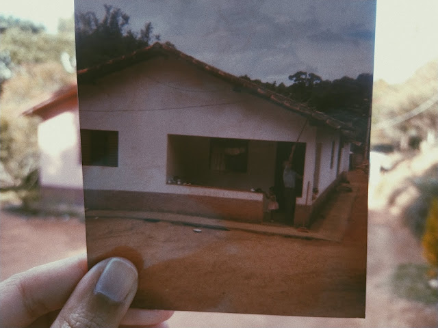 #PraCegoVer: A imagem do começo do texto contém uma pessoa segurando uma foto de uma casa, que se sobrepõe a casa hoje. A imagem da casa antes e depois juntas na mesma fotografia. Passado e presente se entrelaçando.