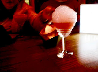 A long stemmed martini glass filled with pink liquid and topped with white bubbles on a large rectangular wooden table on a dark background 