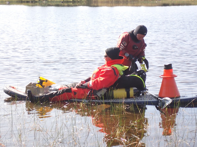 Henkilö istuu pelastautumispuvussa SUP-laudalla, lastattuna sukellusvarusteet ja vesikiikari, kuivapukuinen henkilö taustalla