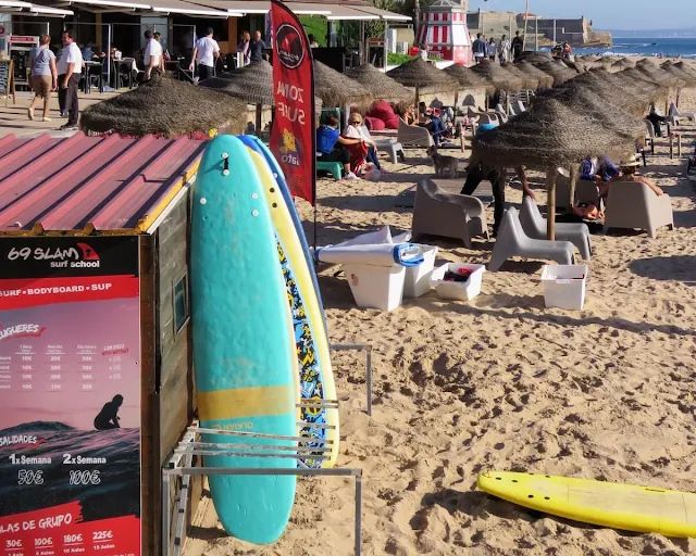 A picture of Carcavelos Beach with turquoise surfboards in the foreground and tiki inspired umbrellas lined up along the beach in the background