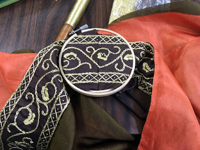 A strip of gold-embroidered brown silk in a tiny wooden hoop, framing the two narrow diamond borders around an undulating oak-leaf vine, with a join in the strip just visible at the vertical center of the hoop. Swathes of green-gold and peach fabric flare from the embroidered strips at either side, and a long wooden bobbin with gleaming gold silk thread rests at upper left on the dark wood table.