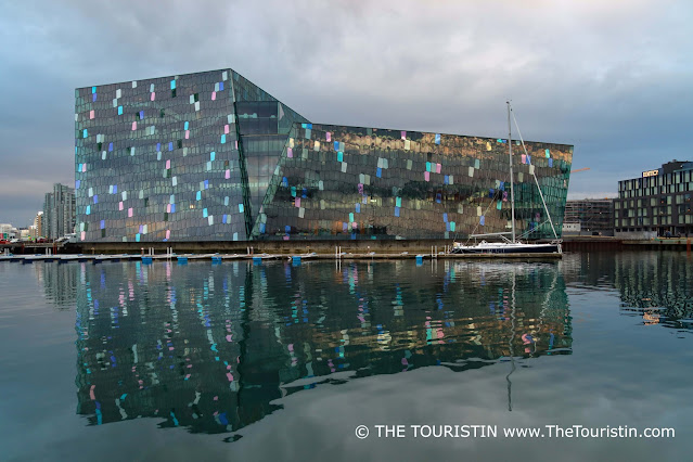 The reflection of a modern building with a coloured glass facade and an unrigged blue sailing boat in calm water.