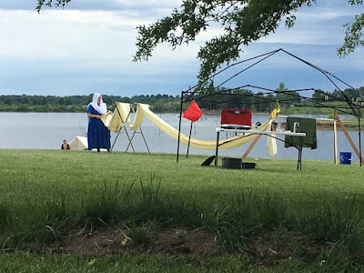 A view across a grassy field to a lake fringed in trees at the far side, with a woman in blue medieval tunic and white veil checking a very long piece of pale-yellow fabric draped over a pair of wooden drying racks, with the black articulated skeleton of a shade fly in the foreground.