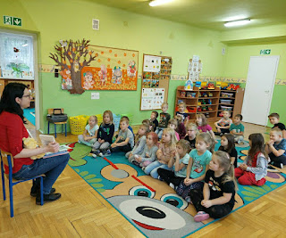 Pani bibliotekarka opowiada o Kubusiu Puchatku, dzieci siedzą na dywanie. Tło: sala przedszkolna.