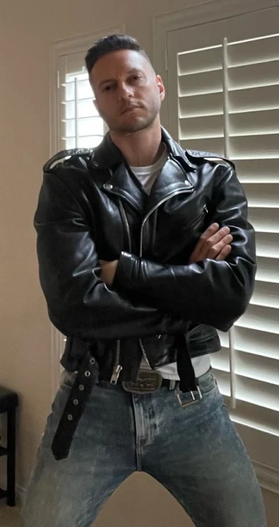 Standing in the living room in front of blinds wearing a black leather biker jacket and blue jeans a handsome man