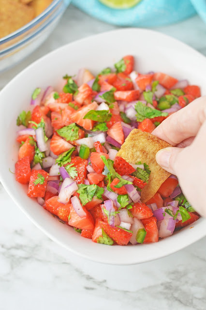 a hand with a chip scooping salsa from a white bowl.