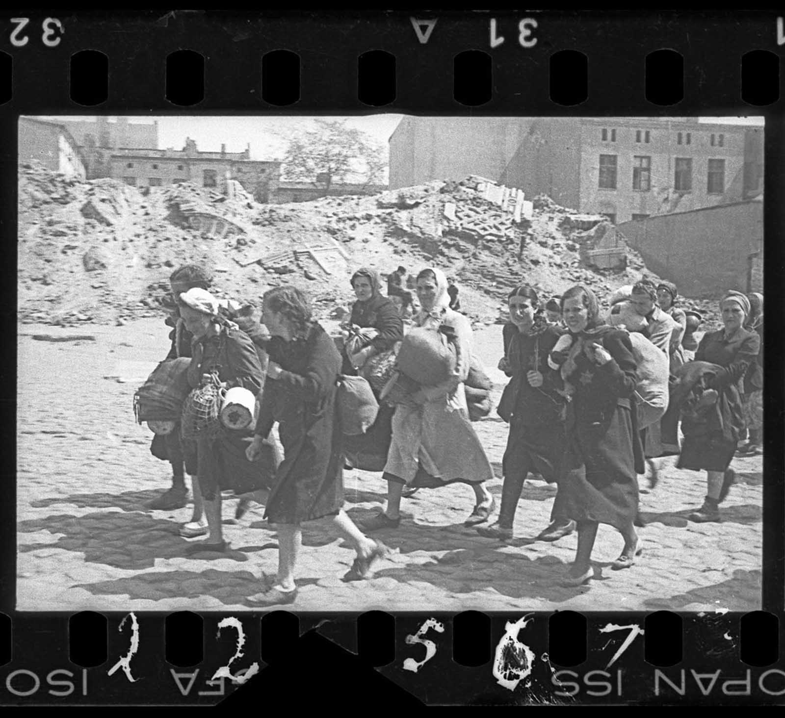 A group of women with sacks and pails, walking past synagogue ruins heading for deportation.