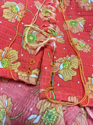 A close photo of the back edges of a coral sundress with large, scattered orange poppy motifs. Two needles threaded with thick variegated orange-and-yellow thread are attached to either side. Three eyelets are worked at the top of both edges, and narrow peach-colored satin ribbon is laced diagonally down the rest of the back.