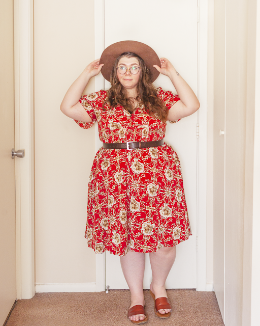 An outfit consisting of brown wide brim hat, a red short sleeve floral midi dress belted with a dark brown belt and brown slide sandals.
