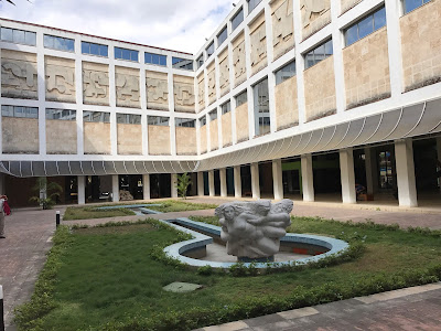 The courtyard of a modern-looking building with relief sculpture on parts of the walls. The courtyard has a large grassy area and a fountain with a sculpture.