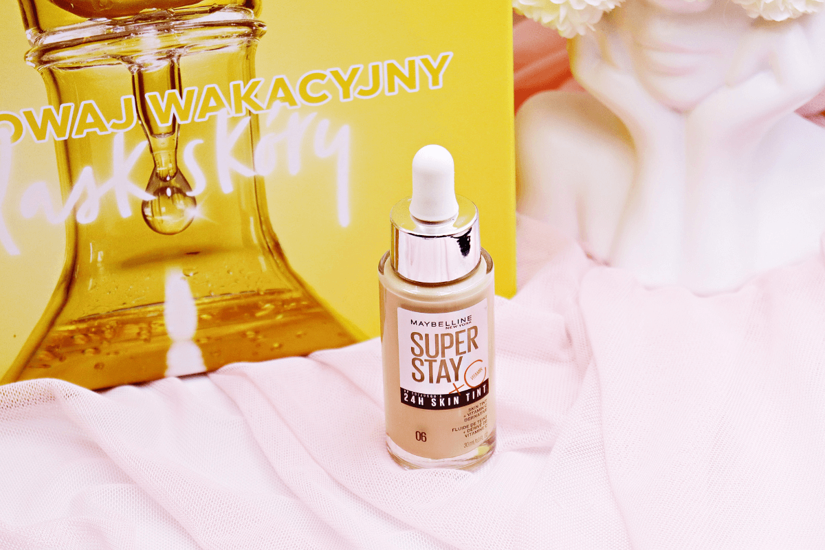 Lekki podkład Maybelline Super Stay 24h Skin Tint na różowym materiałowym tle, z żółtym tłem graficznym w tle promującym wakacyjny blask skóry, wraz z delikatnymi kwiatowymi akcentami, podkreślający jego właściwości długotrwałe i rozświetlające