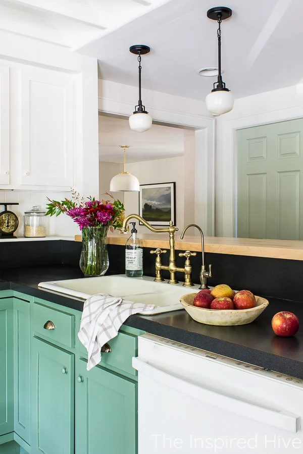 Green kitchen cabinets, black countertop, enamel sink, brass faucet, and schoolhouse lights.