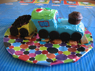 A 3D blue steam engine train cake with a cupcake for a smokestack, followed by a yellow coal car filled with crumbled oreos for coal, and oreos for wheels, on top of graham cracker tracks, sitting on a pizza tray covered with birthday paper (multicolor dots) on a table with a blue Thomas the Tank Engine tablecloth.