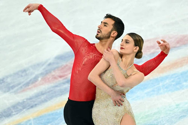 Cizeron e Papadakis em pose inicial de apresentação. Ele veste uma blusa vermelha e estica os braços no ar. Ela está na sua frente, vestindo um vestido dourado