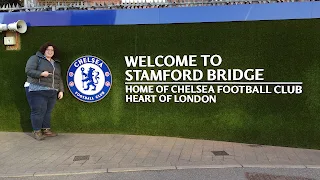 Artifical dark green grass wall with a blue border at the top covered in spikes. On the wall is the symbol for Chealsea Football Club. To the right of the symbol written in white letters it says "Welcome to Stamford Bridge: home of Chealsea Football Club Heart of London." To the left of the Chelsea sign is a woman in a grey coat and blue jeans pointing at it with her right hand and smiling.
