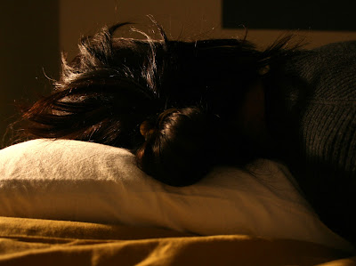 nainen makaa sängyssä pää tyynyllä tummat hiukset sykeröllä selkä kameraan päin