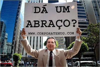 Na imagem vê-se um homem de terno, personagem do documentário O Abraço Corporativo, segurando uma placa onde se lê a frase "Dá um abraço?"."