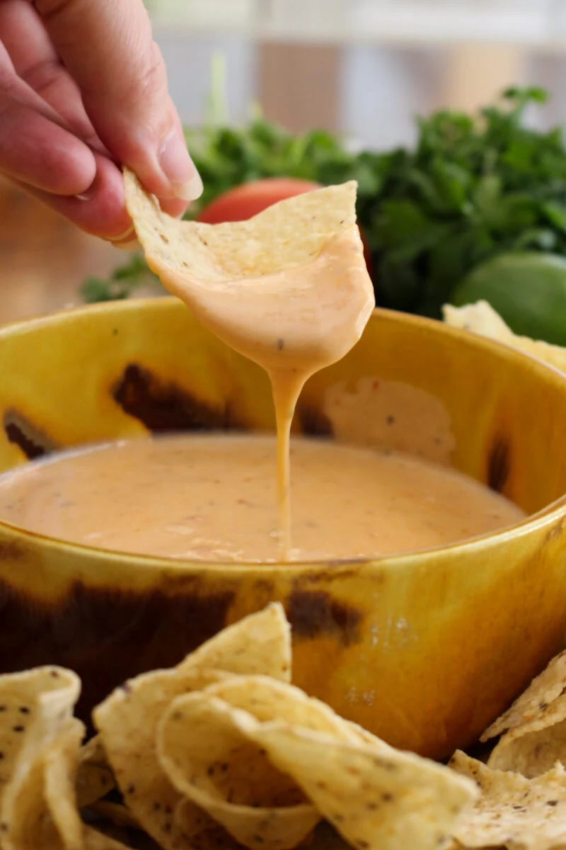 A hand holding a chip that has been dipped in Salsa Con Queso in a yellow bowl.