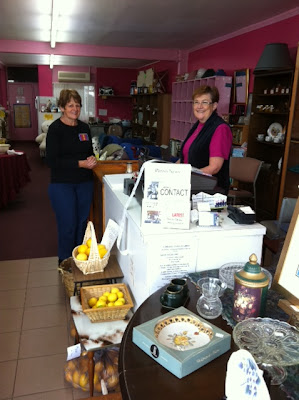 Two smiling ladies standing by the shop counter, with furntiure, decorative items and local fruit