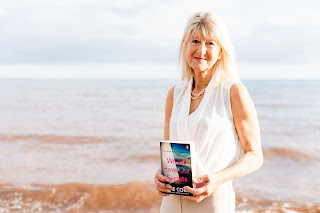 Author photo of Jane Corry. She is a blonde woman standing by the sea holding a copy of her novel We All Have Our Secrets.