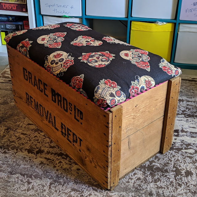 Photo of wooden crate with lid closed placed at an angle to show the side, with fabric covering the lid. The fabric is of skulls and flowers. The front of the crate says "Grade Bros Ltd Removal Dept".